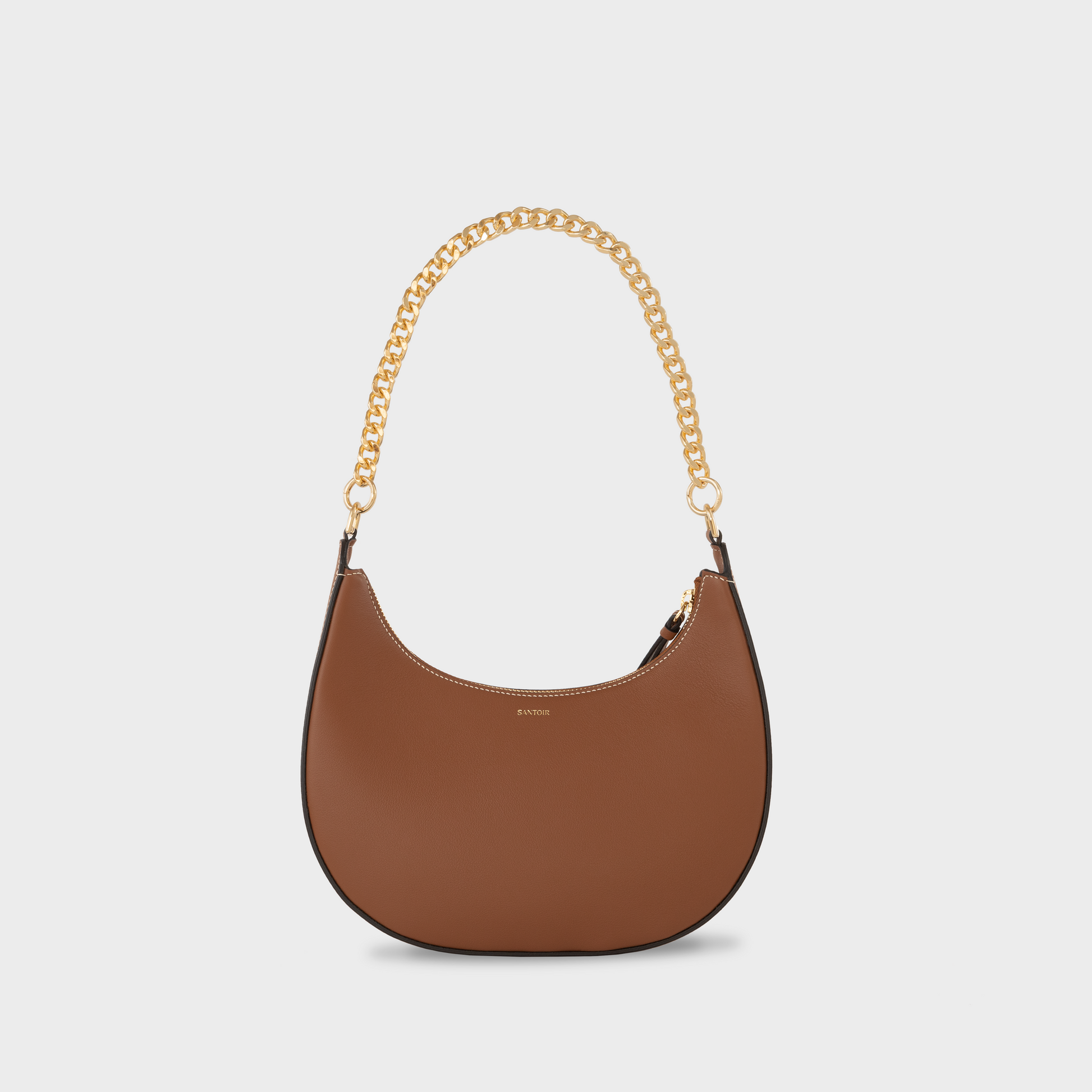 A brown full-grain leather handbag with a smooth finish and a gold-tone chain shoulder strap. Caramel crescent moon handbag with gold chain strap, Santoir Luna bag in smooth warm tan leather, convertible from daytime wear to elegant evening bag, made in Spain.
