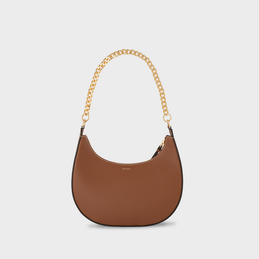 A brown full-grain leather handbag with a smooth finish and a gold-tone chain shoulder strap. Caramel crescent moon handbag with gold chain strap, Santoir Luna bag in smooth warm tan leather, convertible from daytime wear to elegant evening bag, made in Spain.